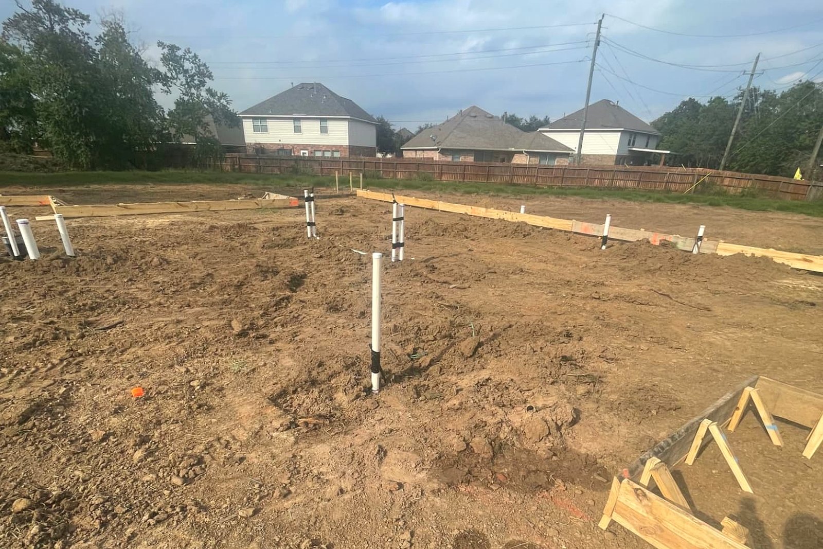 Staked wooden foundation forms and plumbing stub-outs laid out across a graded residential lot